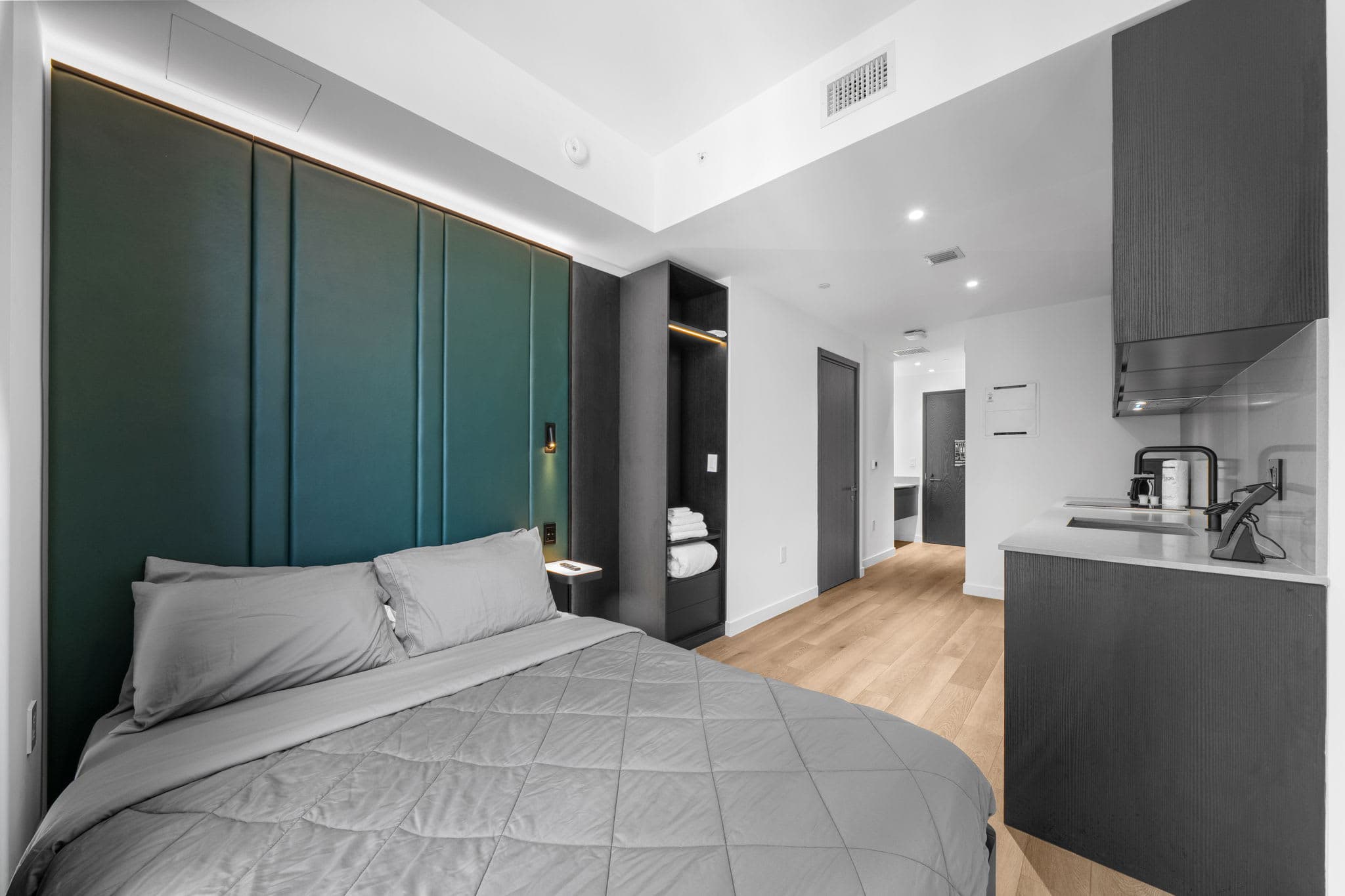 Bed beside a green headboard and compact kitchen.